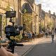 Cinema camera on a gimbal filming a Cotswold village high street with quaint stone buildings and cafe seating, representing narrated property tours and local area videos